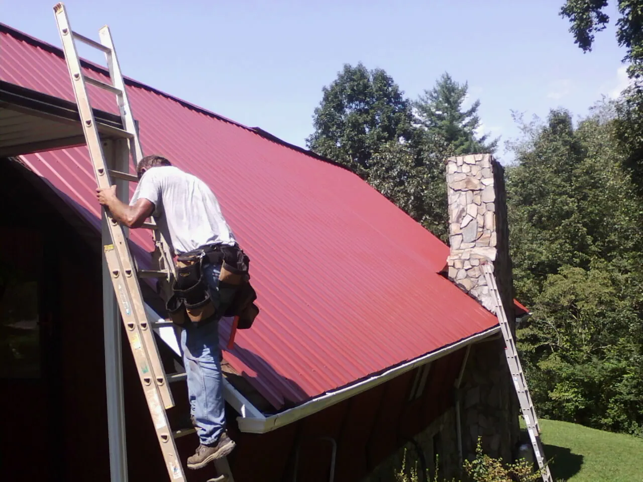 Roofer on a ladder beside a red metal roof and stone chimney — Soffit & Fascia Repair by Roofing Brothers in Herrin